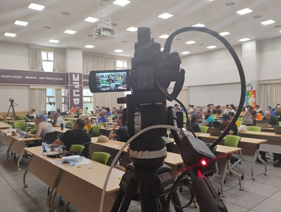 A camera pointing towards a hall of people sitting. A small device next to the camera is lighted red.