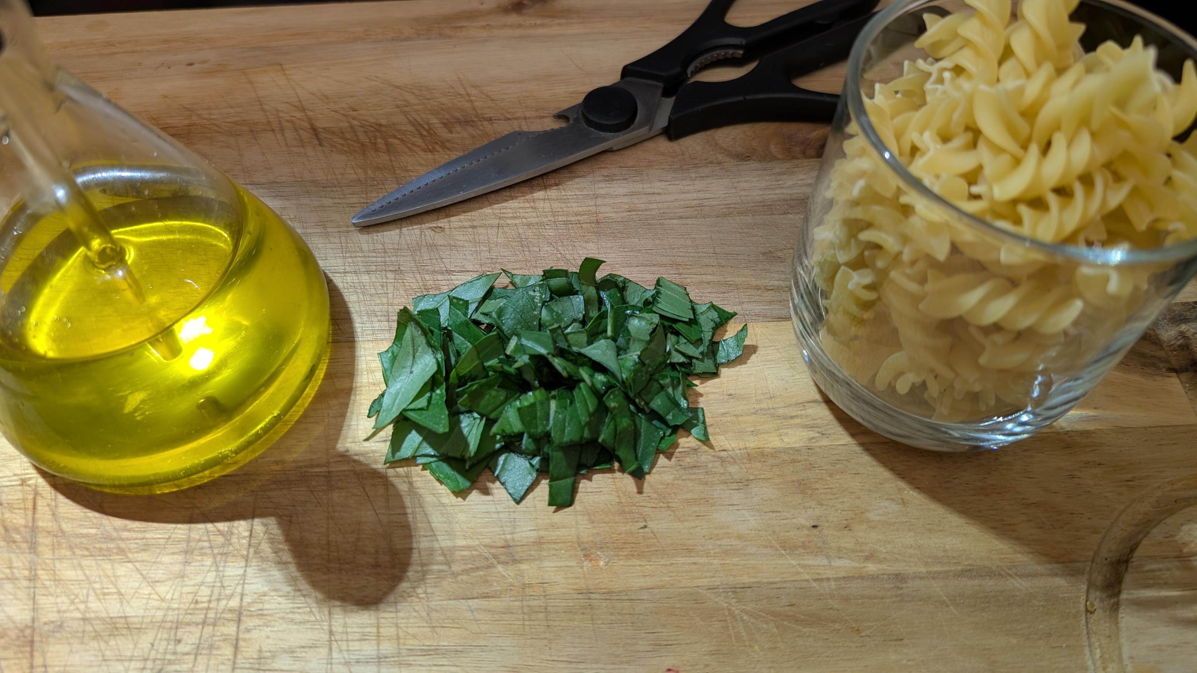 Photo, sur un plateau en acacia, une fiole d'huile d'olive, du basilic coupé en petit morceau, des pâtes dans un verre, et une paire de ciseaux.