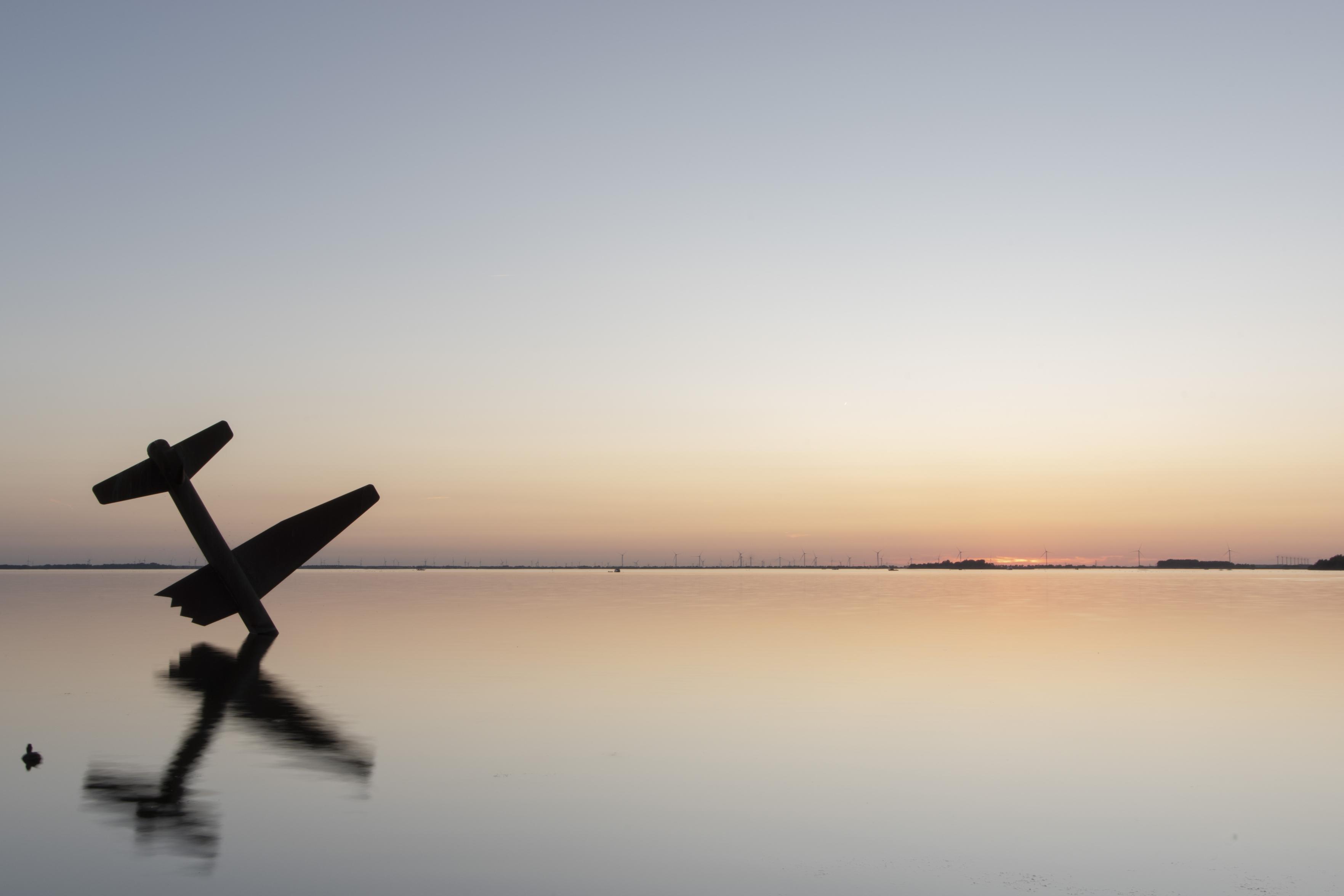 Photo of a monument of a plane with one wing broken flying into water. The monument is in the lower left, the horizon at sunset in the background. In the left is also the silhouette of a duck.