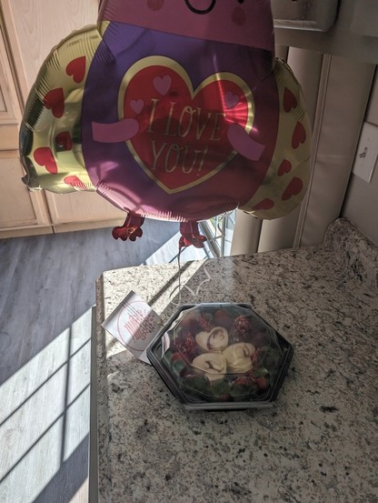 A photo of the Edible Arrangements Valentine's Day gift that my wife got me. There's cheesecake, strawberries and chocolate covered strawberries. There's also a balloon tied to it in the shape of a love bug and it says I Love You.