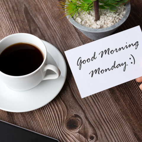 An image that says Good Morning Monday on a white index card. There is a cup of coffee to the left, in a white cup on a white saucer. You can see part of a potted plant in the top right corner and all of this appears to be sitting on a wood table of some sort.