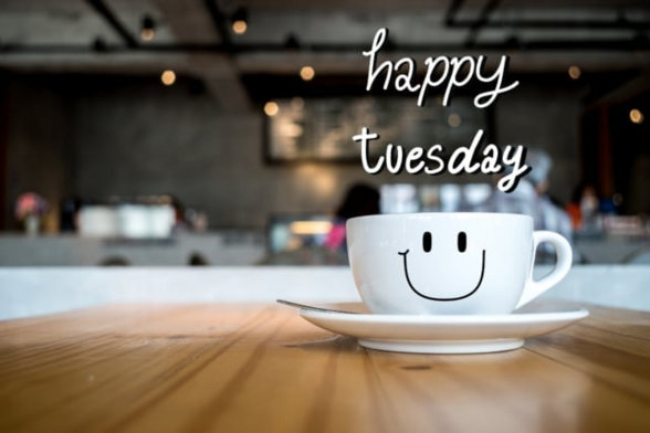 An image that says happy Tuesday in a small, thin white font. You can see a coffee cup on a saucer sitting on a wooden like table. The cup has a smiler face on it in thin black lines. You can see the blurred background of a coffee shop or something similar.