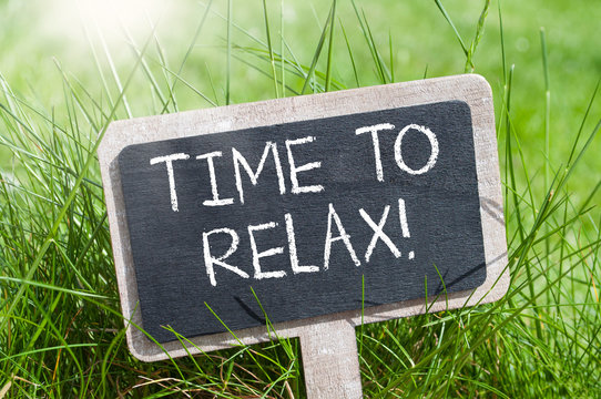 A sign made out of a chalkboard that is in a yard with taller grass. The sign says Time To Relax.