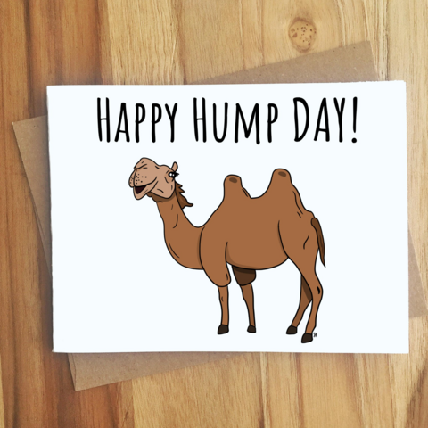 An image with what appears to be a white card with a picture of a camel and the words happy hump day! The card is sitting on a wooden table.