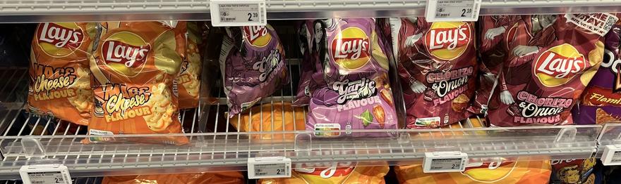 A photo of a grocery store shelf showing the three flavours side by side; orange and yellow for Mac & Cheese, purples for Roasted Garlic & Herbs, and pinkish reds for Chorizo & Onion.