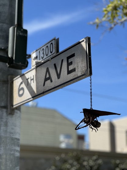 A street sign for 6th Ave with a small metal bug in a hang glider hanging off the end of it from a chain. 