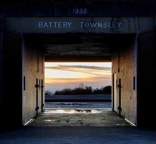 Sunset clouds and silhouetted trees through a concrete tunnel lined with doors and with puddles of water on the ground. A faintly m-visible sign above the near side of the tunnel reads “1938 Battery Townsley”. 