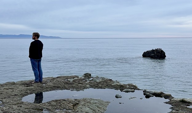 A puddled rocky outcropping over a large body of water stretching to the horizon. A person in a dark sweater stands on the edge of the rock, near the left edge of the photo, looking off into the left distance. On the opposite side of the photo, at the same vertical position as the person and horizontally symmetric, there’s a dark rock sitting in the water.  