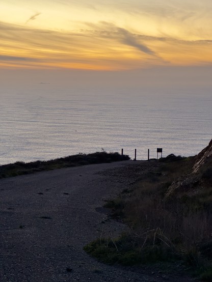 A dark road in bad shape leading to the edge of a cliff, where it stops with a fence. Beyond the cliff is a gray ocean beneath a soft orange and yellow sunset sky. 
