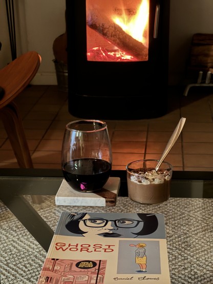 A graphic novel Ghostworld, a glass of red wine, and a pot de crème sitting on a glass coffee table in front of a wood fireplace. 