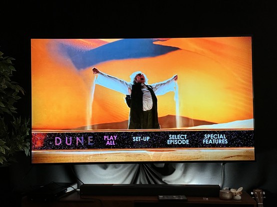 Tv showing the disc menu for DUNE with a person standing with their arms apart, pouring sand out of each, against an orange sand dune. 