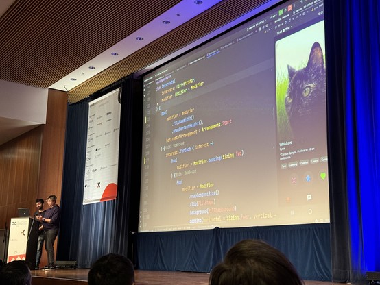 Two people on a stage in front of a large screen showing Android studio with some compose code 