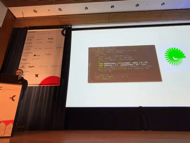 Timo standing on stage beside a large screen showing some shader code and a cool green loading animation. 