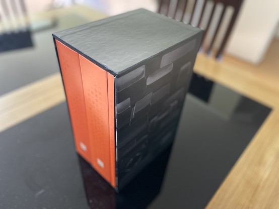 Closeup of the three volumes of Shift Happens (plus leaflet, not visible) inside their sleeve on a desk. The volumes are orange, with subtle symbols on them. The sleeve is dark gray, with key cap shapes without lettering.