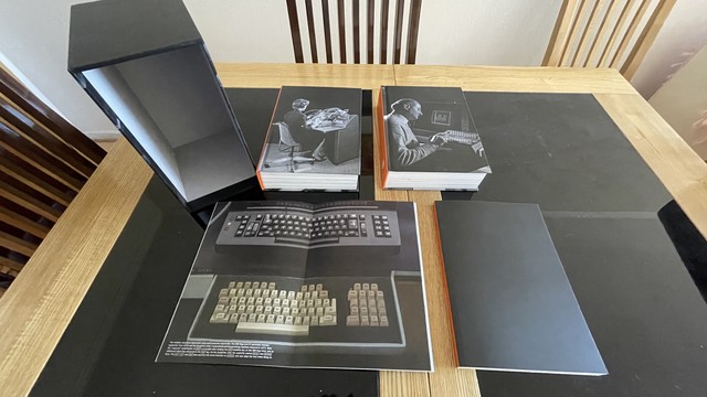 All items from Shift Happens on top of a dark glass and pine table. From left to right, on the top part of the image, we find the book sleeve (empty), Volume One (showing a woman using a teletype on a long exposure; the reflection of the ring shows her hands all over the place), and Volume Two (showing a man typing on a keyboard with two digits, with a. Screen in the background showing some waves of different amplitude). On the bottom part on the left, the central part of the “The Day Return Ba…