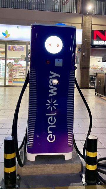 Electric vehicle charger in a strip center. The body is violet, and has “enel way” printed on it rotated 90 degrees counter-clockwise. The charging cables hang from its sides and look like dropped arms.