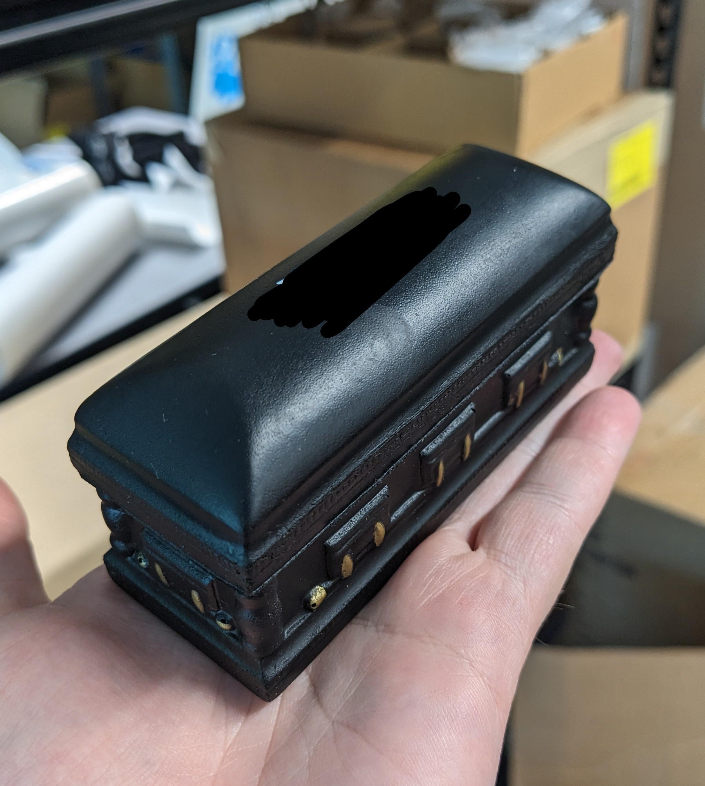 a small foam stress toy in the shape of a casket, black with gold accents, promotional information redacted