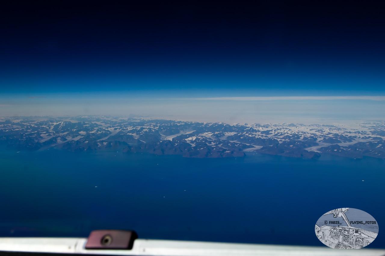 Wide angle view, cockpit windshield wiper in the lower frames deep blue Atlantic right above it leading to the mountains right on the Greenland shore. Iceshield and a few clouds behind it.