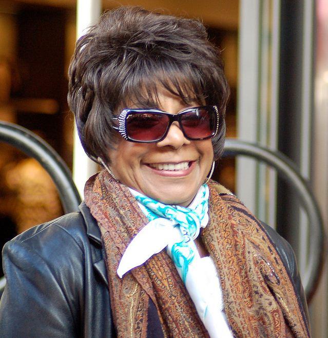 Merry Clayton at a ceremony for Carole King to receive a star on the Hollywood Walk of Fame in 2012. She is wearing a black leather coat with 2 patterned scarves, a pair of large sunglasses with a red-ish tint, and a short feathered hairstyle.
