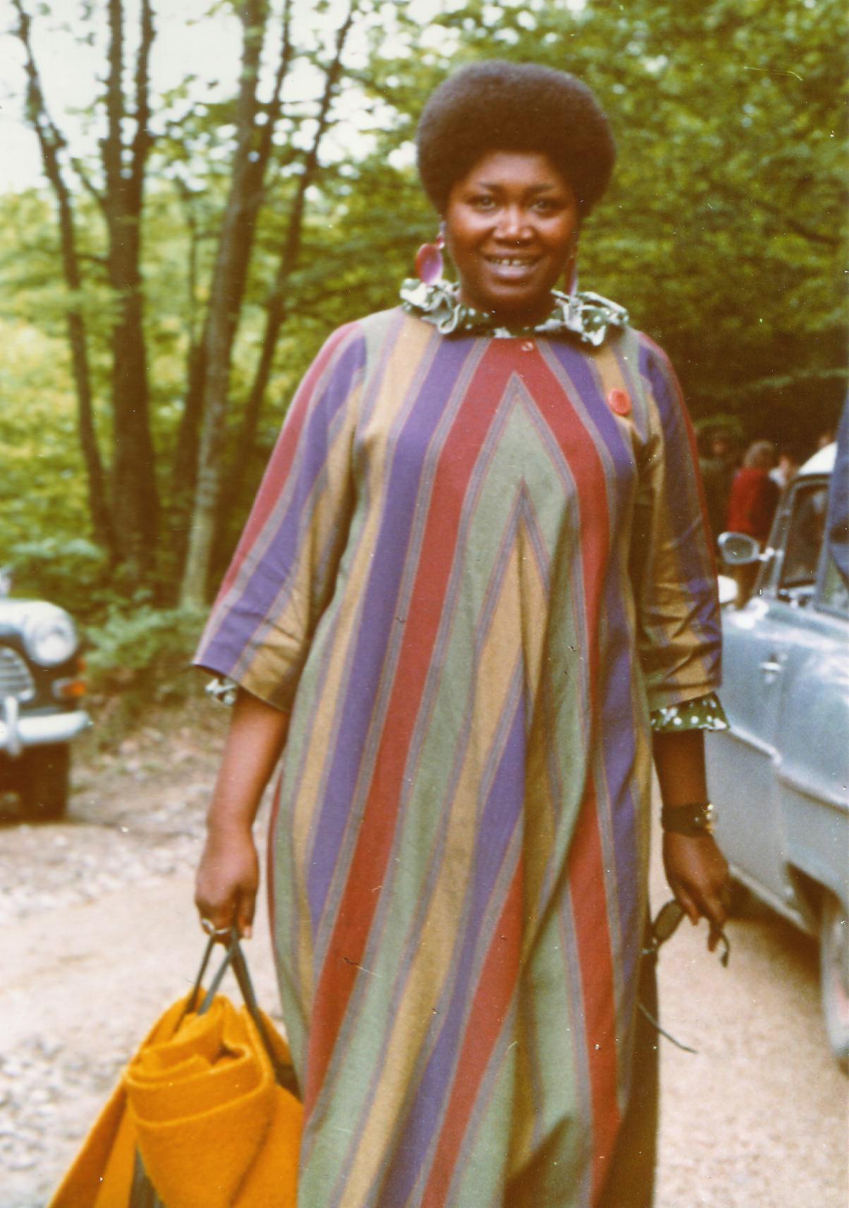 Odetta (Burg Waldeck-Festival 1968, Germany). She is standing near a wooded area. She is wearing a colorful striped muumuu with a ruffled collar. Her hair is styled in an afro and she's smiling while holding a tote bag with a yellow folded blanket in her right hand and sunglasses in her left hand