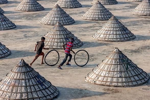 Children play with bicycle tires among large conical piles of rice at a processing mill in Brahmanbaria, Bangladesh