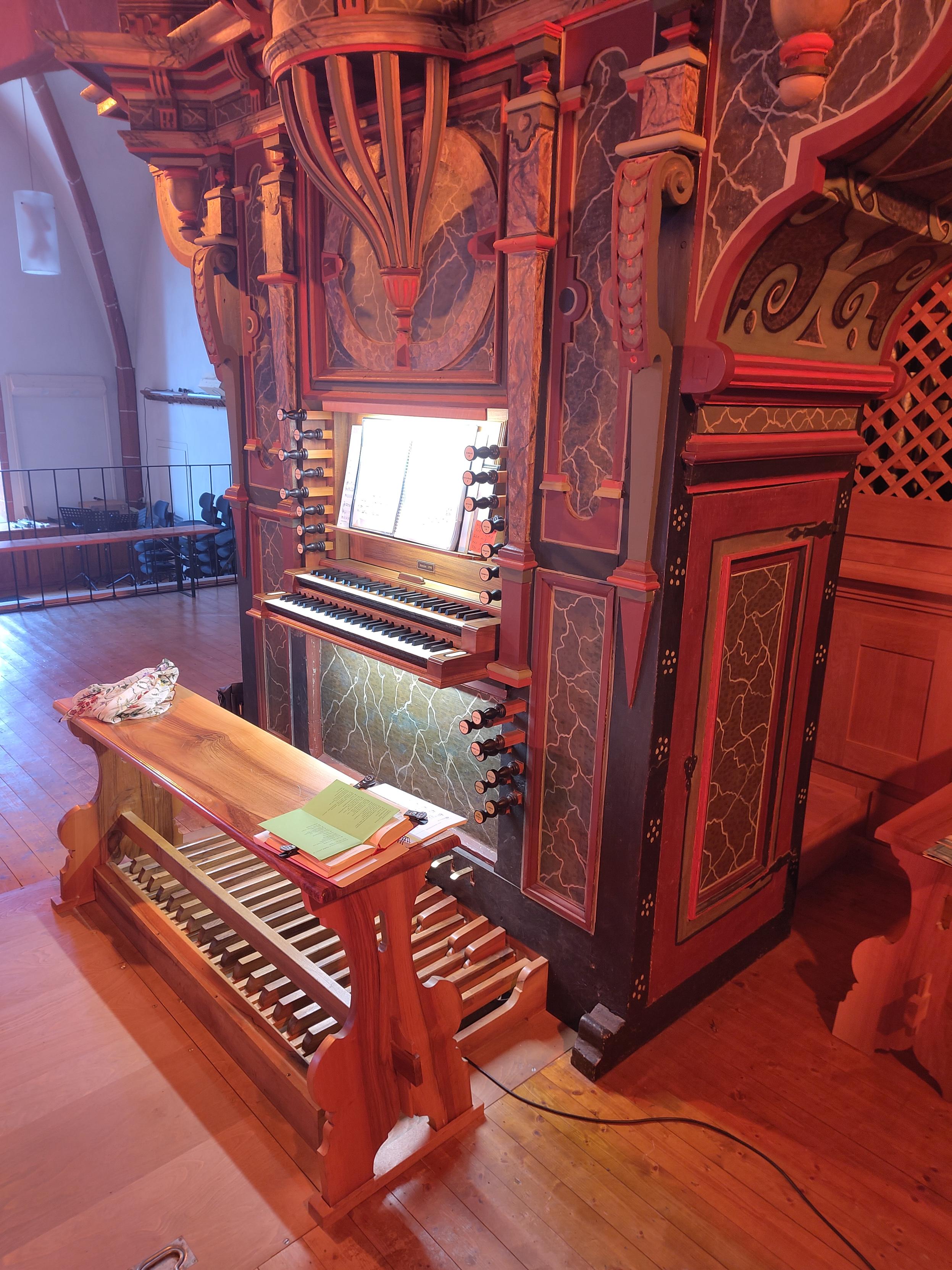Blick auf den zweimanualigen Spieltisch einer Metzler Orgel. Das Gehäuse und der Prospekt sind alt, aus dem Barock, die Orgel selbst ist von 1990. In Rottönen, reichlich verziert.