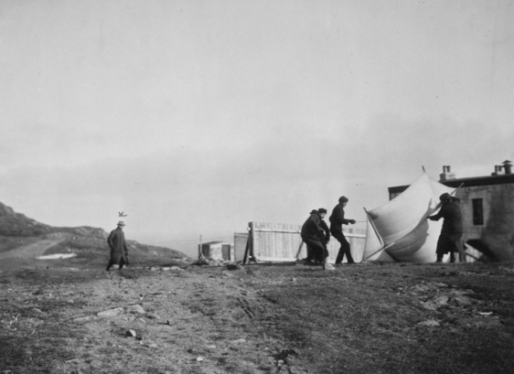Several men in dark suits are trying to raise a flag on a relatively barren landscape in this black and white photo.