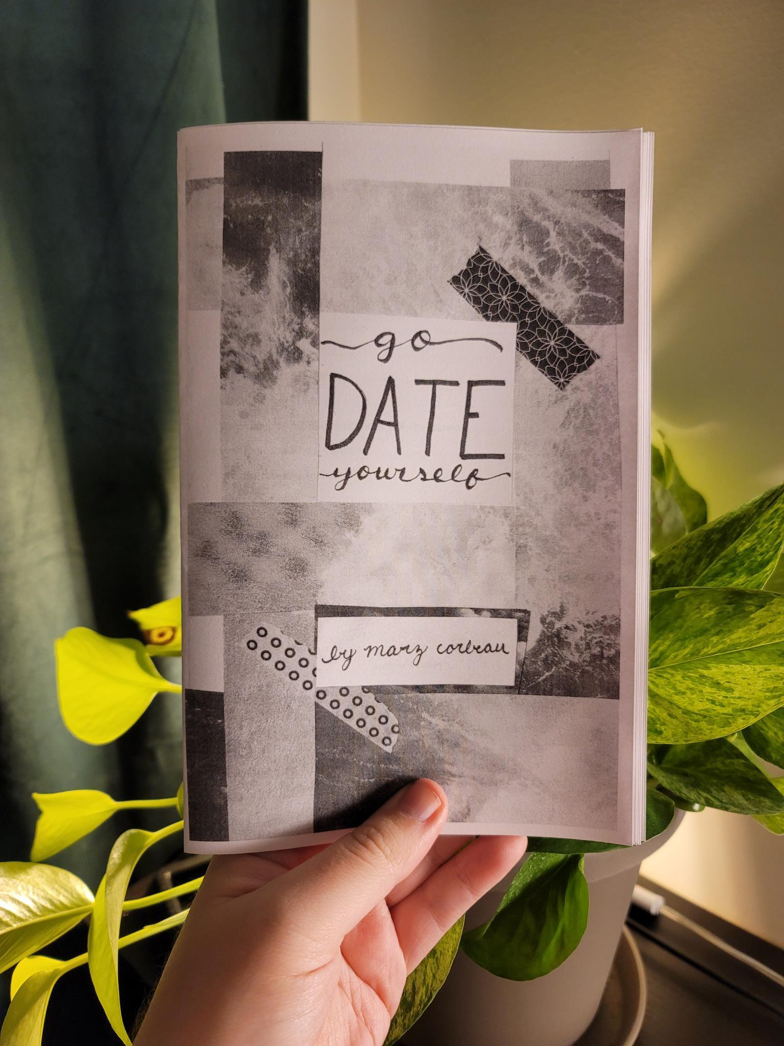 A white hand holding a black and white copy of a zine titled "Go Date Yourself" in front of a backlit pothos plant. 