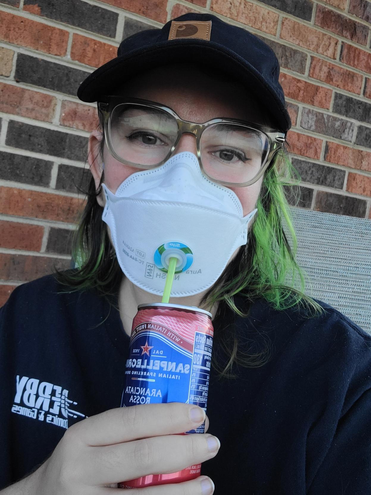 White person with black and green hair and dark green eyes in a black cap, green glasses, and black shirt in front of a brick wall. There's a white valve in the mask with a blue and green cover on the edges of it. Through the valve is a green straw that goes into a tall thin red soda can. They smile from behind their mask and look happy in the evening light.