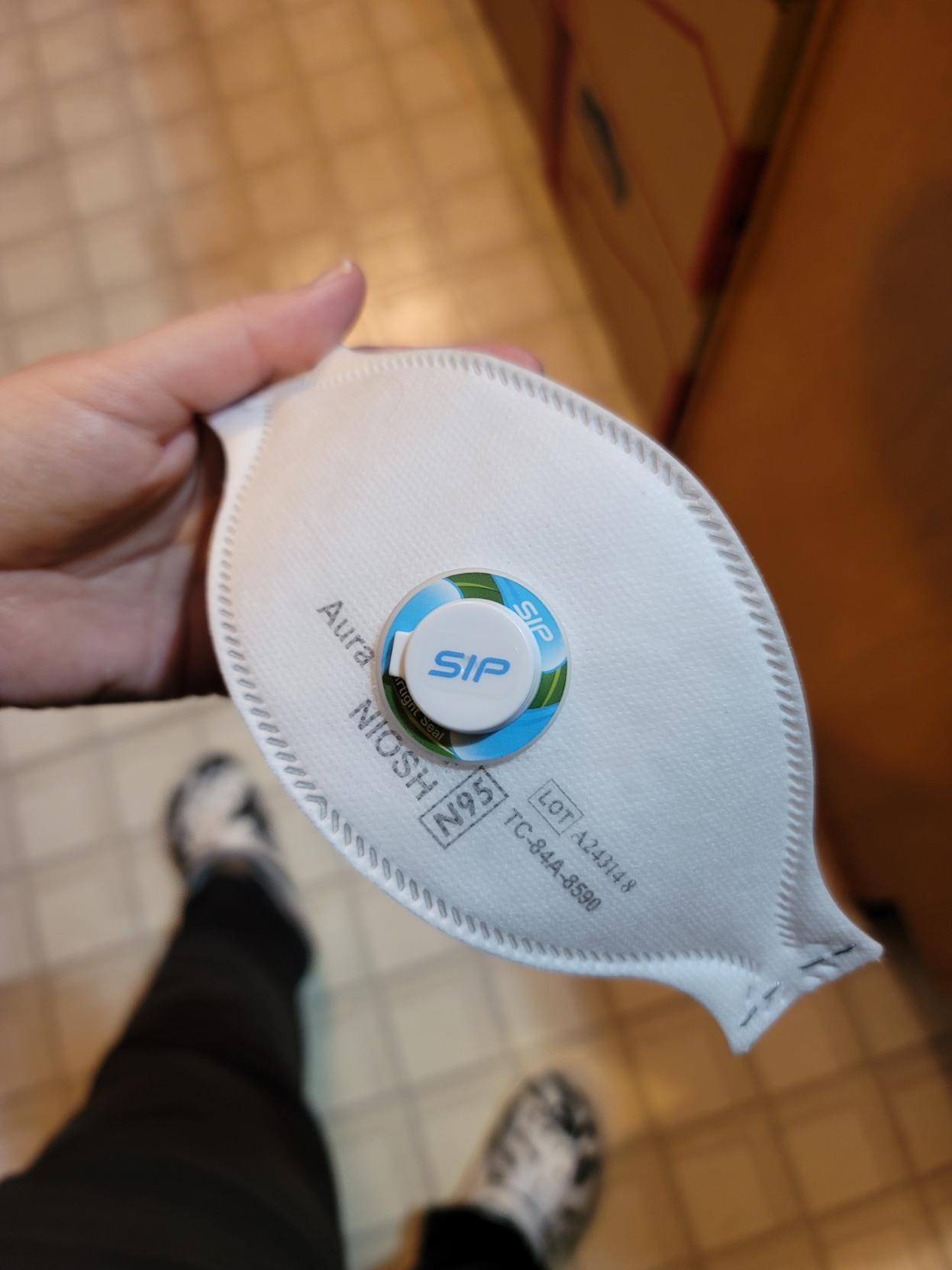 A white hand holds the 3M Aura mask with sip valve through the center under indoor florescent light for closer examination.