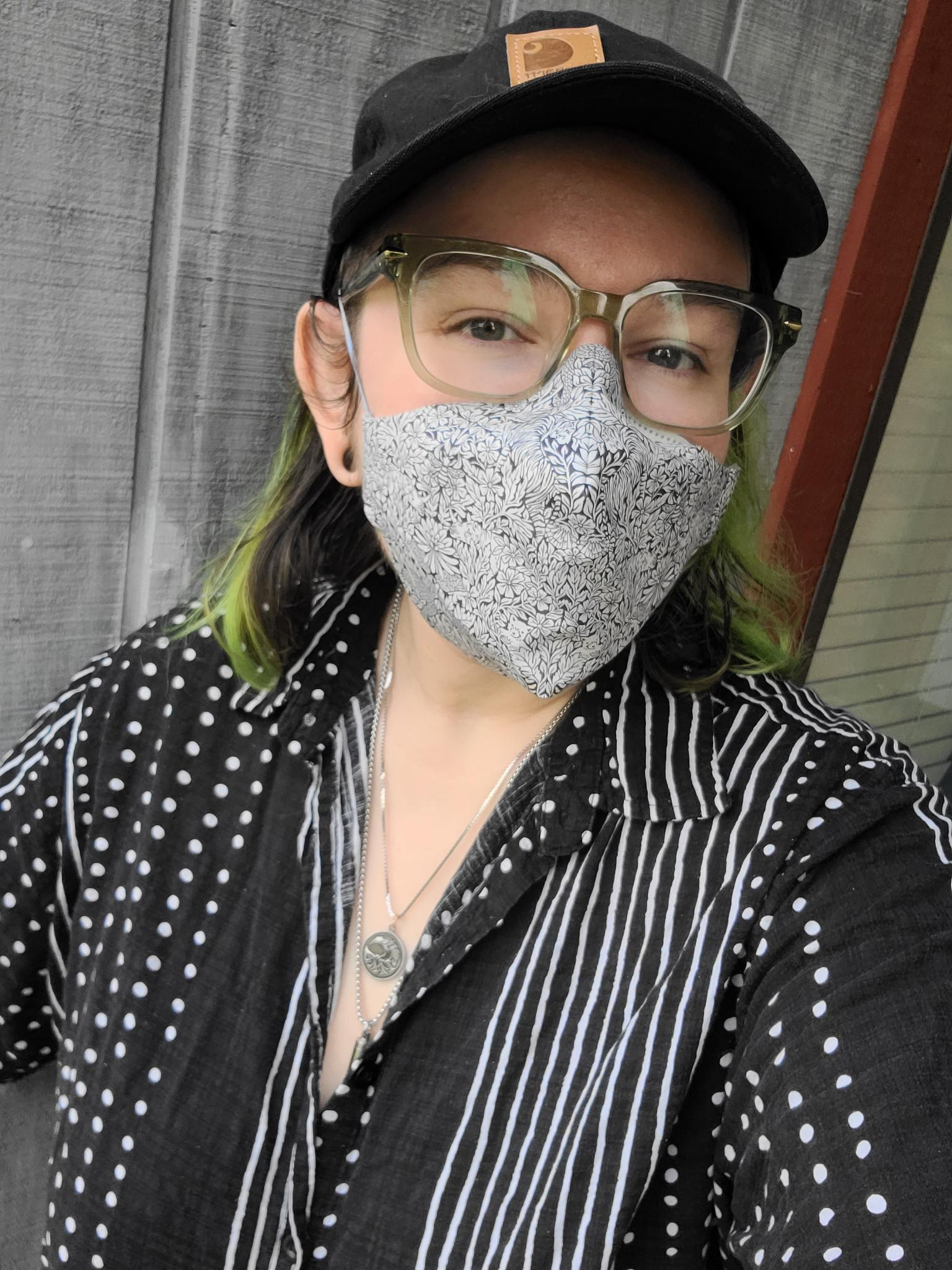 Photo of a white person with black and green hair, black cap, green glasses, and black and white button up shirt showing off a cloth cover for their 3M Aura respirator. The cloth cover has a fine floral print in white and navy. 