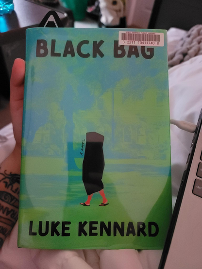 Photo of a white hand holding a book entitled Black Bag by Luke Kennard. It has a blue and green photo of a college campus with a body bag with legs - specifically in flip flops - illustrated on it. The text is bold and black at the top and bottom of the cover. 