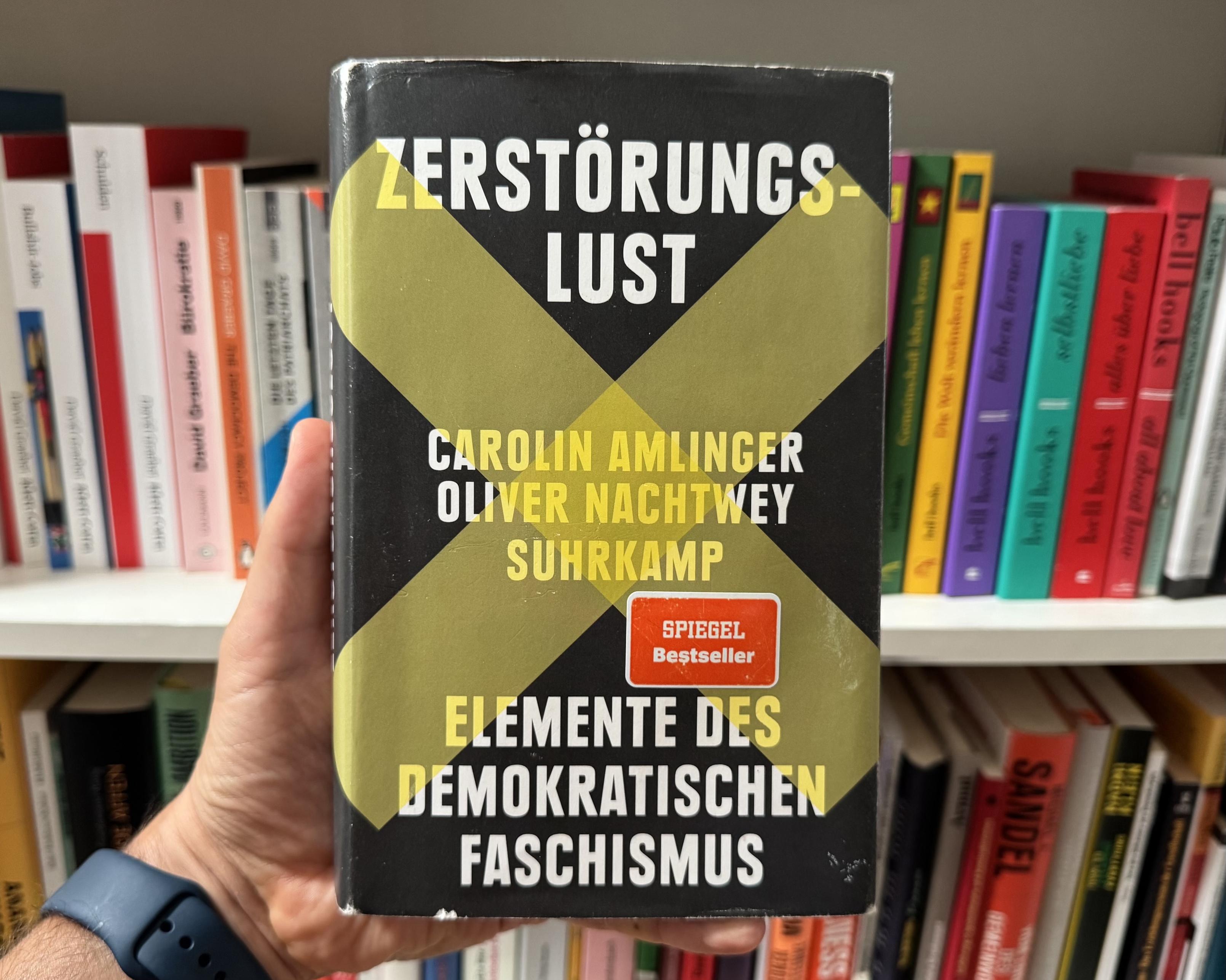 (M)Eine Hand hält das Buch „Zerstörungslust“ von Carolin Amlinger und Oliver Nachtwey (erschienen im Suhrkamp Verlag) in die Kamera. Im Hintergrund ist ein unscharfes Bücherregal zu sehen. Das Cover ist schwarz und wird von einem großen, olivgrünen oder dunkelgelben „X“ dominiert, das sich über die gesamte Fläche erstreckt. Der Titel und die Autorennamen sind in serifenloser Großschrift gedruckt, wobei die Farben zwischen Weiß und Gelb kontrastieren: Oben steht „ZERSTÖRUNGSLUST“, mittig folgen die Autorennamen sowie der Verlagsname „SUHRKAMP“, und unten befindet sich der Untertitel „ELEMENTE DES DEMOKRATISCHEN FASCHISMUS“. Auf der rechten Seite, leicht unterhalb der Mitte, ist zudem ein kleiner, rechteckiger, orangefarbener Aufkleber mit der weißen Aufschrift „SPIEGEL Bestseller“ aufgebracht.