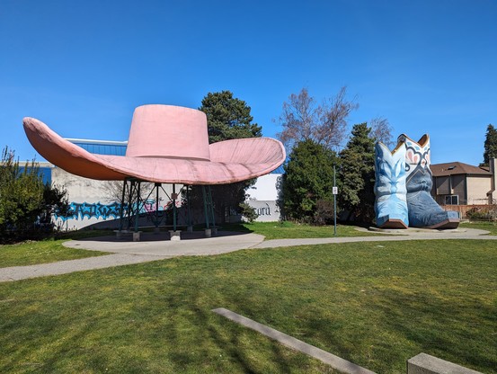 A two story tall pair of cowboy boots, and a forty foot wide hat, in a small green park.

The sky is deep blue overhead.

This picture focuses more on the hat — brick red almost pink, with a wide brim — and less on the boots.