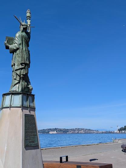 The Statue of Liberty, on Alki Beach, seen from one-quarter profile (slightly behind Lady Liberty, on her left).

In the distance you can see the Seattle Space Needle across Elliott Bay.

The sky and bay are a deep blue.
