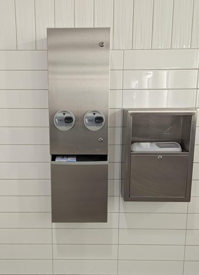 A stainless steel dispenser on a tile wall. 

It appears to have two eyes (buttons, marked FREE) looking audience right, and the dispenser real looks like a mouth. 

If you zoom in you can see that the thing in the dispenser tray is a box of menstrual pads