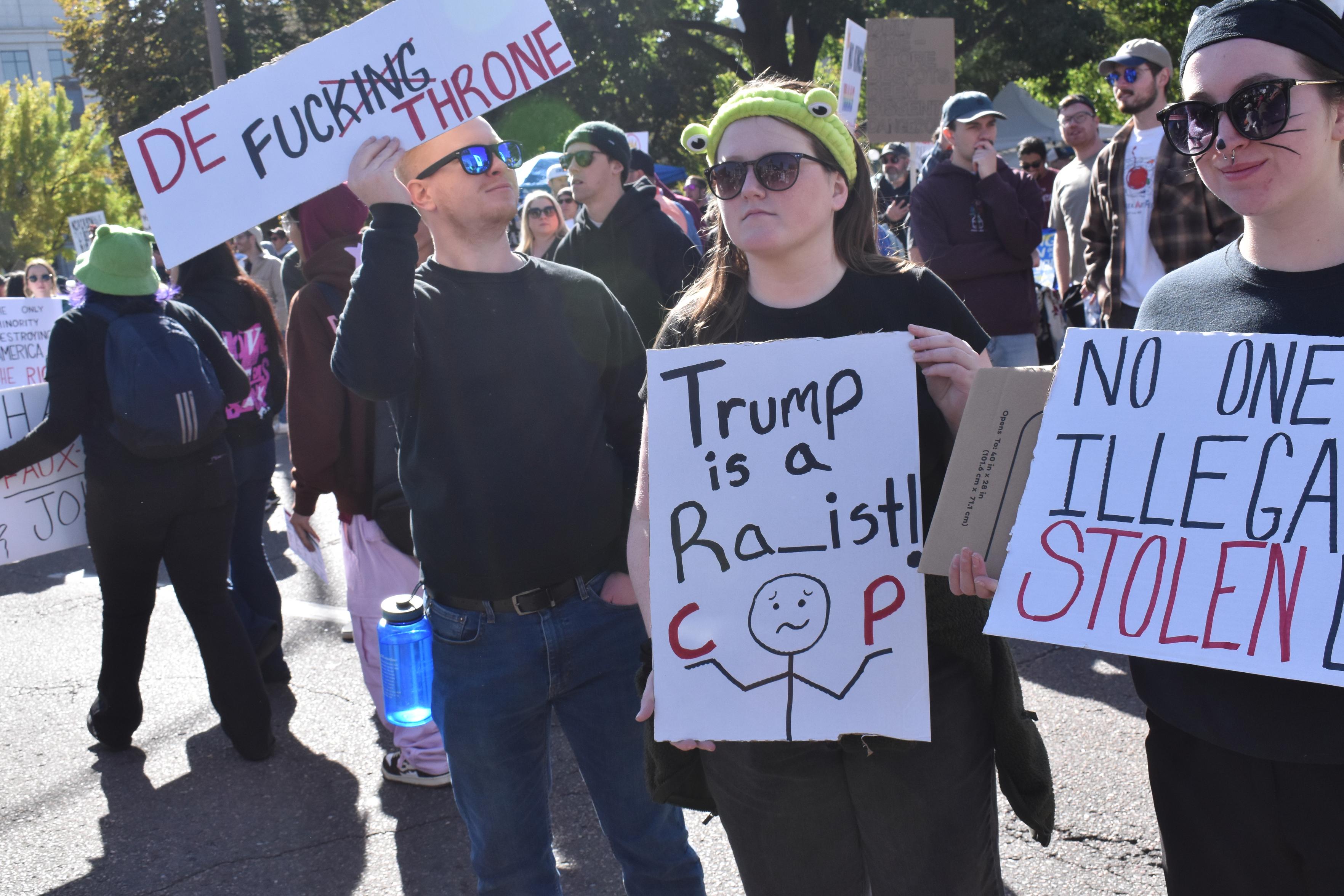 Two women with hand held signs dressed in black. Sign says "Trump is a RA_ist!" Then a stick figure with a confused look has a C in one hand and a P in the other.