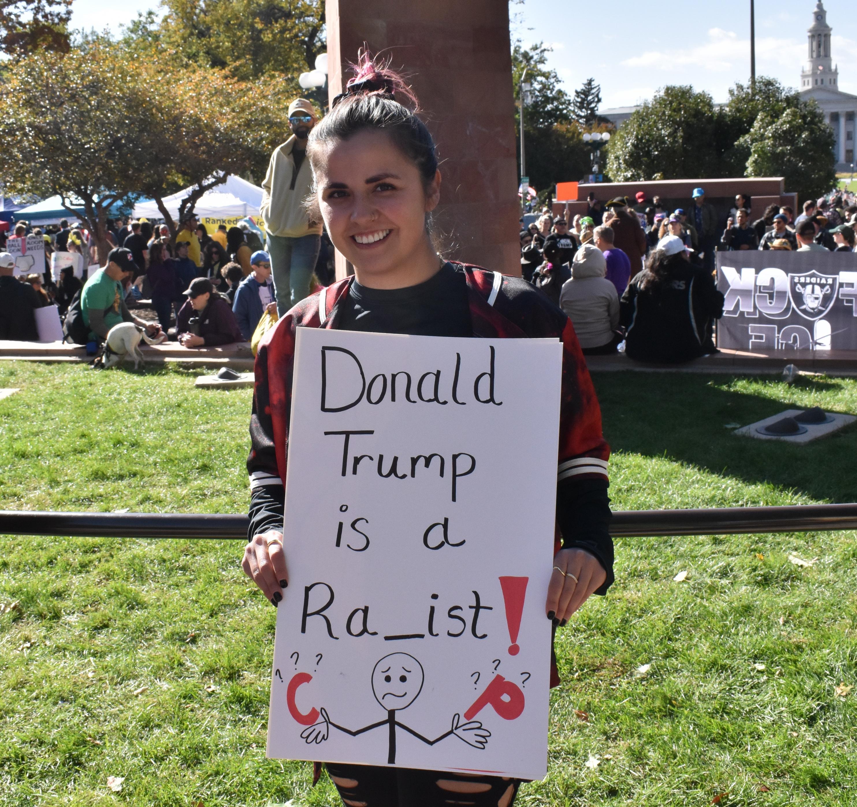 Young woman with dark hair bulled back is smiling holding a sign that reads, "Donald Trump is a RA_ist!" beneath that is a stick figure with a confused look. In one hand is a red C and in the other is a red P. Both leftters have smaller question marks around them.