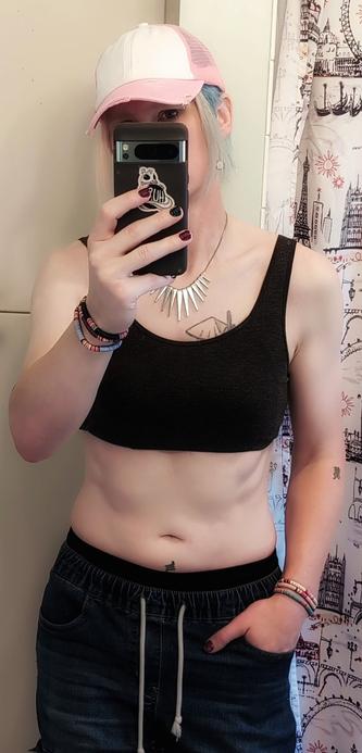 Alice stands confidently before the mirror, capturing a moment that blends tomboy charm with raw sensuality. The pink and white baseball cap casts a playful shadow over their eyes, hinting at the mischievous smile hidden beneath. Their cropped, dark heathered gray tank top clings to their chest, the absence of a bra leaving little to the imagination as it reveals the soft curves of their toned form. Cargo jeans hang low on their hips, the drawstrings dangling loosely, inviting easy access. The waistband of black cotton panties peeks out provocatively above the jeans, hinting at what's below. Alice’s hands—one tucked confidently into a pocket, the other holding their phone—show off alternating glittery purple and teal nails, each color catching the light in a flirtatious glimmer. This is Alice in full tomboy mode, effortlessly mixing rugged and sexy in a way that says fuck around and find out...or find out if we'll fuck around.