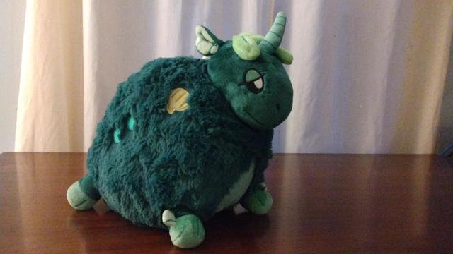 A green plush, part horse and part sea creature, with a single horn, and bits of seaweed. They have heavy-lidded eyes and a smile.