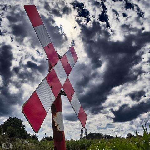 Backlighted crossbuck to indicate a level railway crossing. Behind it a dramatic sky with the sun peeping through.