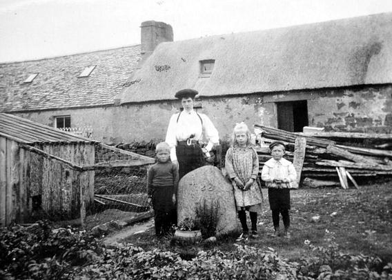 An antique photograph in grainy black and white of a rural location. A well dressed family of a woman and three children, two boys and a girl all stand in back of a weathered stone in the ground, a small boulder which is now called the 'Janet Horne' memorial stone. Legend has it this spot represents the location where the 'last witch' was killed in Scotland. A long stone building and piles of lumber are in the background.