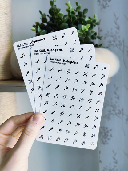hand holding up three white sticker sheets with tiny black icons showing different weapons like swords, blunt weapons and arrows