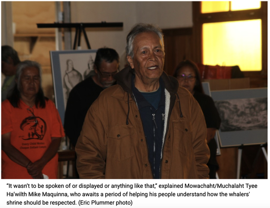 A dark skinned First Nations man stands and speaks. He is wearing a heavy brown jacket. Some people are standing behind him. There are also some drawings or paintings. They are standing in hall of the church at Yoquot.

The caption says:
"It wasn't to be spoken of or displayed or anything like that," explained Mowachaht/Muchalaht Tyee Ha'wilth Mike Maquinna, who awaits a period of helping his people understand how the whalers'
shrine should be respected. (Eric Plummer photo)