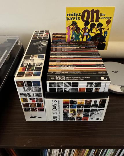 photograph of a CD boxset, Miles Davis - The Complete Columbia Album Collection. the set comprises 53 albums pressed on CD in carton sleeves with the artwork of the original record releases, in a carton box with a hardbound booklet. the box is between a turntable and a portable CD player. the cover of Miles Davis’ On the Corner is on display.