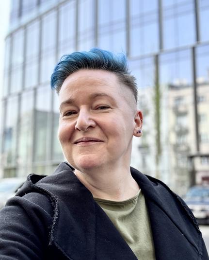 Osoba zadowolona prosto od fryzjera, z krótko obciętą czupryną w kolorze niebieskim i wygolonymi bokami, w czarnej bluzie, stoi na tle jakiegoś wieżowca w którym odbija się niebo i domy naprzeciwko. Ma przymrużone oczy i uśmiecha się nieznacznie lecz dumnie
