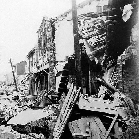 Black and white photo of two-story storefront buildings with sections of brick walls and timber fallen in front of them, extending down the street. A telephone pole leans in the background.