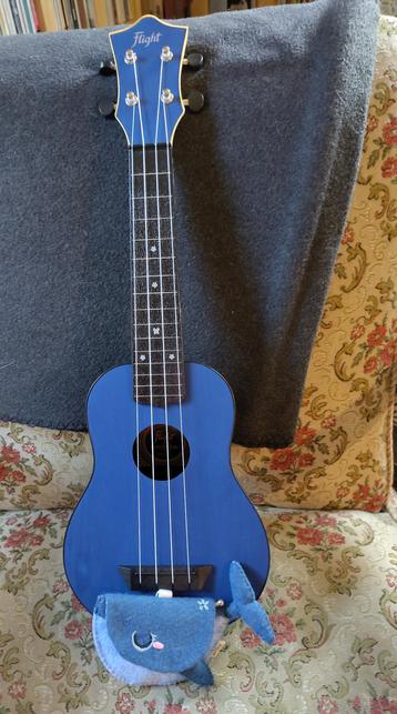A blue soprano ukulele leans against an easy chair. At its bottom, there's a felted pouch in form of a whale in two shades of blue. It's about the size of the palm of my hand.