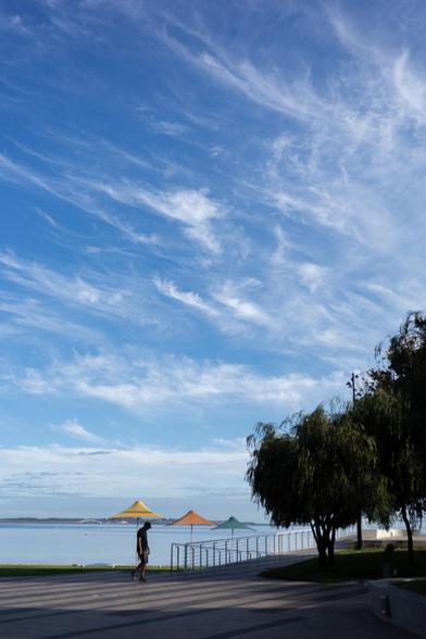 A person walking along a promenade near the water under a blue sky with wispy clouds. There are colourful shade structures in the distance and a large tree on the right side.