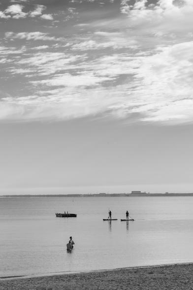 A black and white photo of a calm beach scene. In the distance, two people are paddleboarding on the water. In the foreground, a man stands in the shallow water while another person is partially submerged nearby. The sky is partly cloudy, and there is a distant view of a shoreline with industrial buildings.